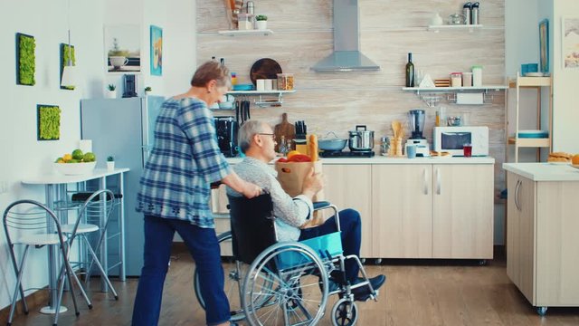 Old woman pushing invalid senior husband in wheelchair after arriving with grocery paper bag from supermarket. Mature people with fresh vegetables for cooking breakfast.