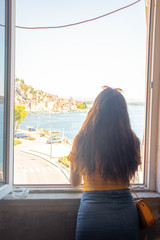 Brunette with long hair seen from behind, standing on a window frame looking into the distance. Outline of the town of Sibenik in the distance. Traveling Croatia and the adriatic sea