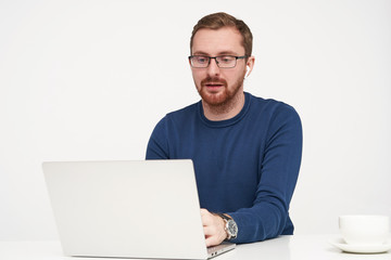 Indoor shot of young fair-haired man in glasses looking seriously at his laptop while typing text on keyboard, being concentrated on his work while posing isolated over white background