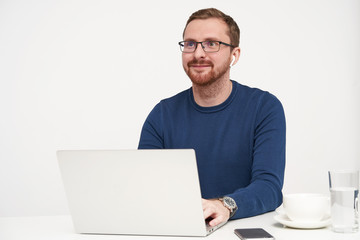 Horizontal shot of young pleased bearded fair-haired man looking positively ahead with lovely smile while keeping hands on keyboard, posing over white background