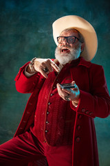 Pointing on phone. Modern stylish Santa Claus in red fashionable suit and cowboy's hat on dark background. Looks like a rockstar. New Year and Christmas eve, celebration, holidays, winter's mood