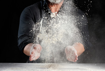 chef in black uniform sprinkles white wheat flour in different directions, product scatters dust