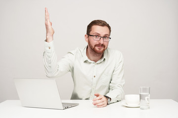 Studio shot of young bearded guy dressed in formal clothes keeping his lips folded while looking at camera and raising up hand, sitting over white background