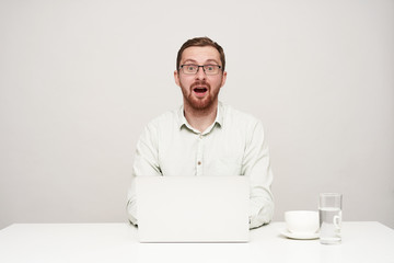 Bemused young pretty bearded man dressed in white shirt looking surprisedly at camera with wide mouth opened while typing text on his laptop, isolated over white background