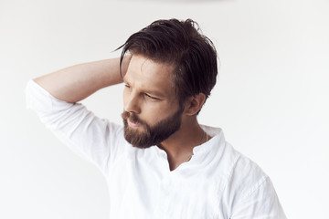 high key portrait photo on white cyclorama of a handsome bearded man with brown hair and eyes, he is wearing a white linen shirt, he holds his hand behind his head and looks away