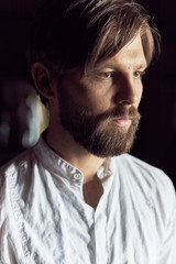 low key portrait of a handsome bearded man with brown hair and  eyes, he is dressed in a white linen shirt and looks away