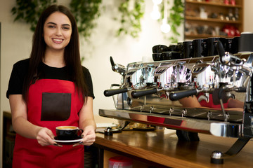Fototapeta na wymiar Portrait of beautiful waitress with aroma cappuccino in hands.Cheerful barista holding cup of tasty americano with milk.Smiling young professional standing near coffee machine while looking at camera