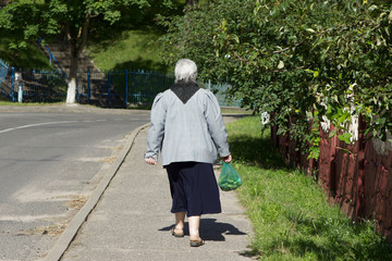 A poor lonely old woman, a pensioner, walks along the city sidewalk with a package of vegetables in the summer. Back view.