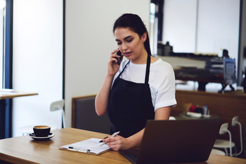 Obraz premium Attractive waitress talking on smartphone and making audit products at cafe indoors.
