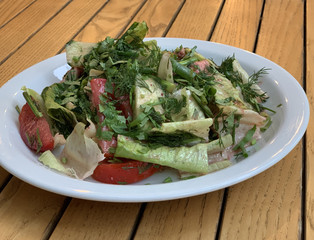 Vegetable salad of cucumbers, tomatoes and herbs on a white plate. Finely chopped vegetables in a salad bowl, on a wooden table. Concept: healthy food, fresh vegetables, vitamins.