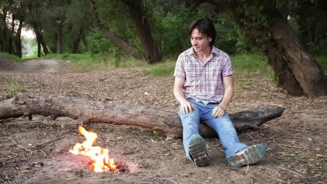 A man sits on a log near a fire, enjoys nature, fights off mosquitoes. Mosquitoes attack a man. A person treats the skin with a spray from mosquitoes, sprays the anti mosquito in the air.