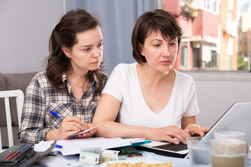 Obraz premium Two smiling young females looking financial documents with laptop in home interior. High quality photo