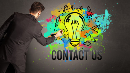 businessman drawing colorful light bulb with CONTACT US inscription on textured concrete wall, new business idea concept