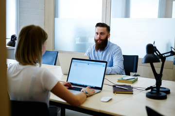 Back view of female professional programmer browsing information on modern laptop with mock up screen connected to wireless internet and discussing ideas for startup project with colleague in office