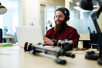 Fototapeta na wymiar Positive male creating music using application on laptop computer and enjoying made composition via headphones.Bearded man in cool hat listening favorite playlist sitting in coworking