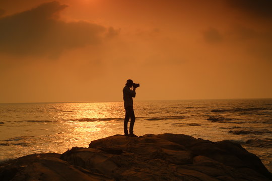 A tourist stand on rock and sea water facing sunset and clicking picture background wallpaper. 