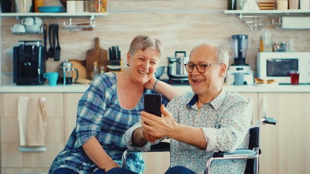 Smiling senior woman and her disabled husband in wheelchair using smartphone in kitchen. Paralysied handicapped old elderly man using modern communication techonolgy.