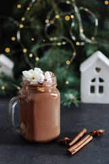 Hot chocolate in a glass mason jar with marshmallows and cinnamon and anise spice on black table surrounded with fir tree, Christmas lights and decorations.