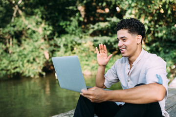 Afro and black man working with his laptop or computer on the street near the river, making a video call with his friends.