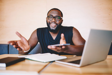 Cheerful african american guy gesturing and holding smartphone satisfied with prosperous freelance business, happy dark skinned male looking at camera smiling completed task of distance job