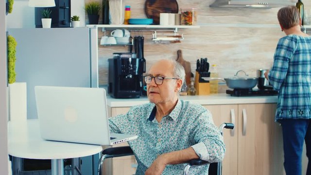 Disabled senior man in wheelchair working on laptop in kitchen while wife is cooking. Handicapped businessman. disabilty entrepreneur paralysis for elderly retired man