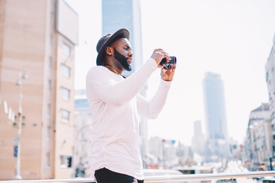 Skilled African American Photographer Using Vintage Camera For Taking Pictures On Urban Setting, Dark Skinned Hipster Guy In Trendy Outfit Making Photos During Vacation Trip While Sightseeing