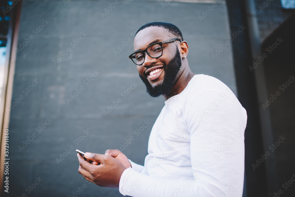Poster portrait of cheerful african american hipster guy in eyewear sending messages on cellular phone sati