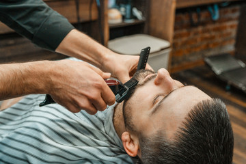 Beauty shop for men. Shaving a beard in a barbershop. Barber cuts his beard with a razor and clipper. close up Brutal haircuts. Hairdresser equipment. Selective focus.
