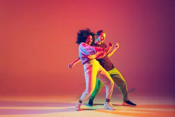 Poster Drive in motion. Stylish man and woman dancing hip-hop in bright clothes on green background at dance hall in neon light. Youth culture, movement, style and fashion, action. Fashionable portrait. © master1305