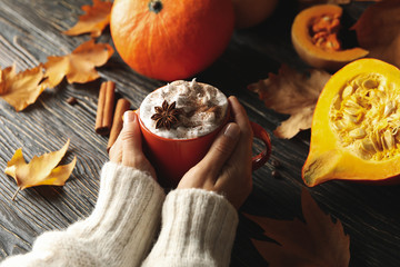 Female hands hold cup of pumpkin latte on wooden background with pumpkin, cinnamon and leaves