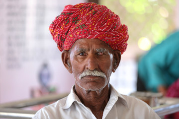 Old Indian man in long white mustache and wearing red turban suffering from eye problem. Cataract in eye. Loss of vision. Damaged one eye. Eye disease. 