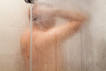 middle aged man takes a shower standing under a jet of water in a shower stall behind a transparent fogged glass door in the bathroom