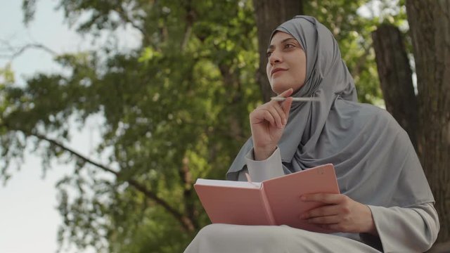 Low angle view of young Muslim woman wearing grey hijab is sitting under the tree, thinking and then writing in pink notebook