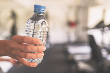 Cropped. Girl hand with bottle still water in the fitness gym opposite the sport equipment and  jogging simulators. Resting time. Health concept.