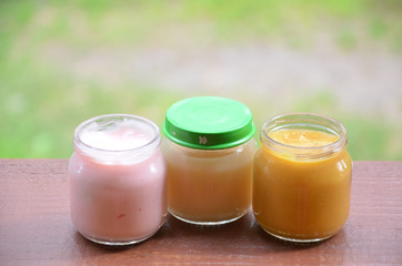 Three jars of baby food on the background of foliage. Pink light yellow orange color