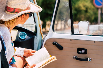 Travel people concept in summer or spring season - caucasian woman read a book sitting inside a retro van and looking at the road - young girl passenger inside a vehicle enjoy the trip