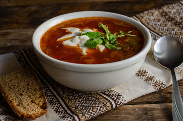 Borscht in a white bowl, bread, metal spoon, kitchen folk. Napkin in the home kitchen on a wooden table.