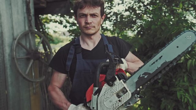Worker in the garden. Young adult man stands in the summer garden with running hand saw