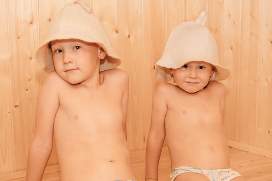 Two little boy, with bath hats on their heads and turned into white sheets looking on each other, beauty and health concept, indoor closeup portrait.