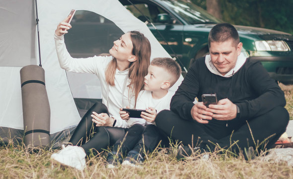 Family sitting in nature near the tent mom takes a selfie with her son and dad sits on the phone