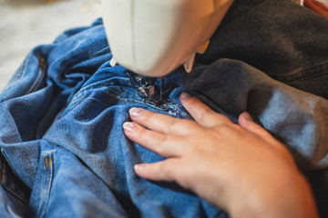 Process of repairing and sewing the jeans at home, close up of denim on the sewing machine, hemming, tailoring and stitching jeans cloth and dress, with the hand of female dressmaker in background