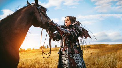 Girl in medieval knight's armor with a horse against the sunset fields background