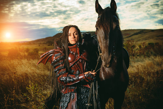 Girl in medieval knight's armor with a horse against the sunset fields background