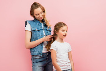 young woman in denim vest and jeans braiding hair of daughter isolated on pink