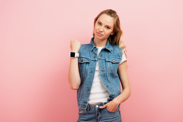 young woman in denim clothes showing smartwatch on wrist while standing with hand in pocket isolated on pink