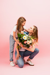 girl in jeans presenting bouquet of flowers to mother and hugging her on pink