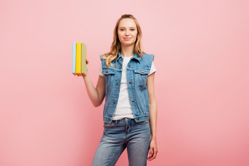 Naklejka premium young woman in denim vest and jeans holding books isolated on pink