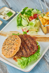 Delicious grilled  burger on white plate on wooden table.  with sauces, french fries and salads.