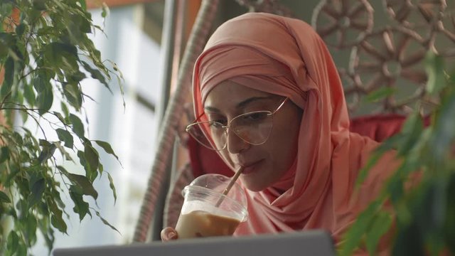 Close up of young smiling mixed-race woman wearing hijab and eyeglasses is sitting in restaurant, drinking milkshake and looking at laptop screen