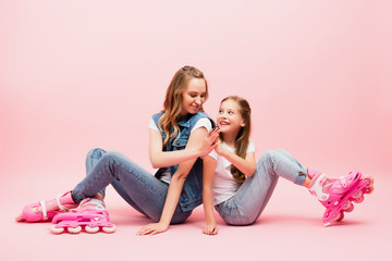 mother and daughter in denim clothes sitting on floor in roller skates and giving high five on pink